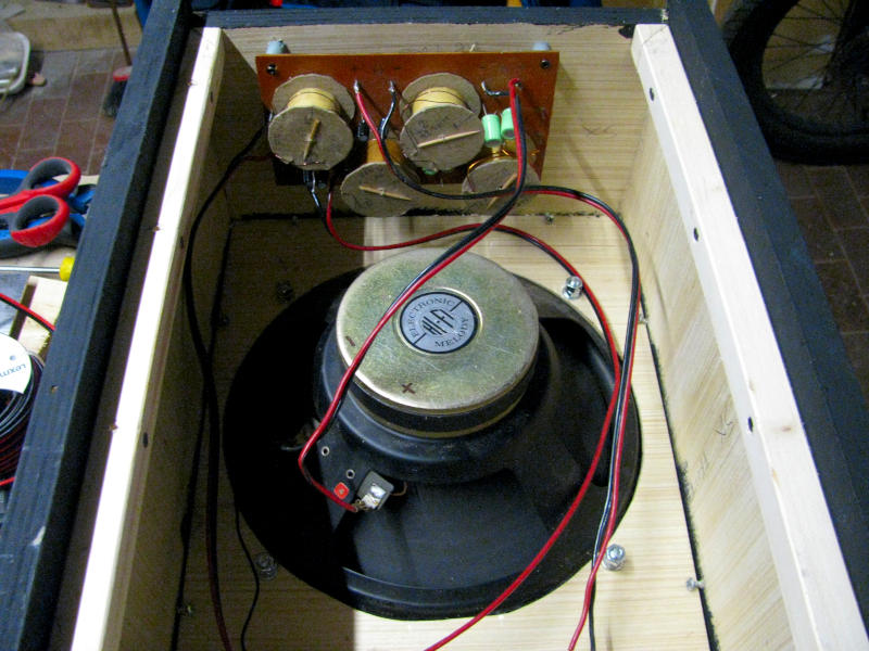 Interno della cassa acustica karaoke con crossover passivo a tre vie e cablaggio degli altoparlanti prima della chiusura Vista dell'interno della cassa acustica per karaoke prima della chiusura del pannello posteriore. In alto è visibile il crossover passivo a tre vie autocostruito montato su basetta in bachelite con bobine di induttanza e condensatori. Al centro il magnete del woofer a sospensione pneumatica da 25cm con i cavi di collegamento rosso e nero. Il profilato ad L nero è visibile lungo il bordo interno della cassa.
