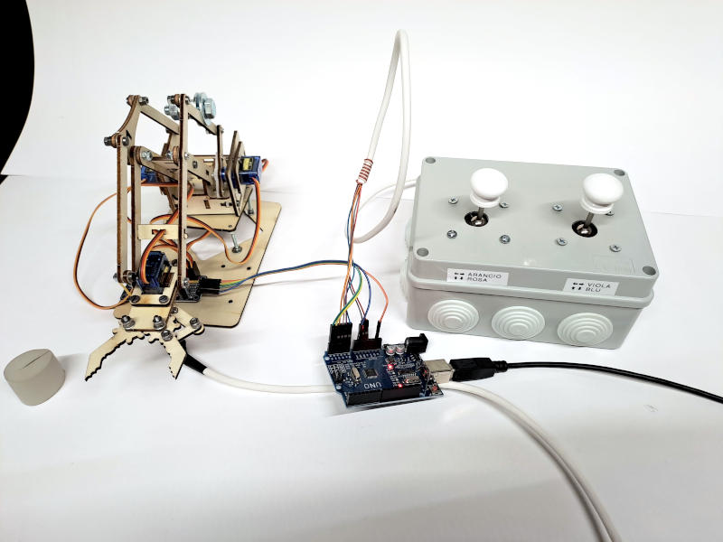 Vista d'insieme del sistema di controllo completo. Da sinistra: il braccio robotico 4DOF in legno con la scheda PCA9685 alla base, Arduino Uno al centro con i connettori Dupont sugli ingressi analogici e I2C, e il controller a due joystick nella scatola di derivazione stagna a destra. Il cavo multipolare bianco collega il controller ad Arduino, mentre il cavo USB fornisce alimentazione e connessione al computer